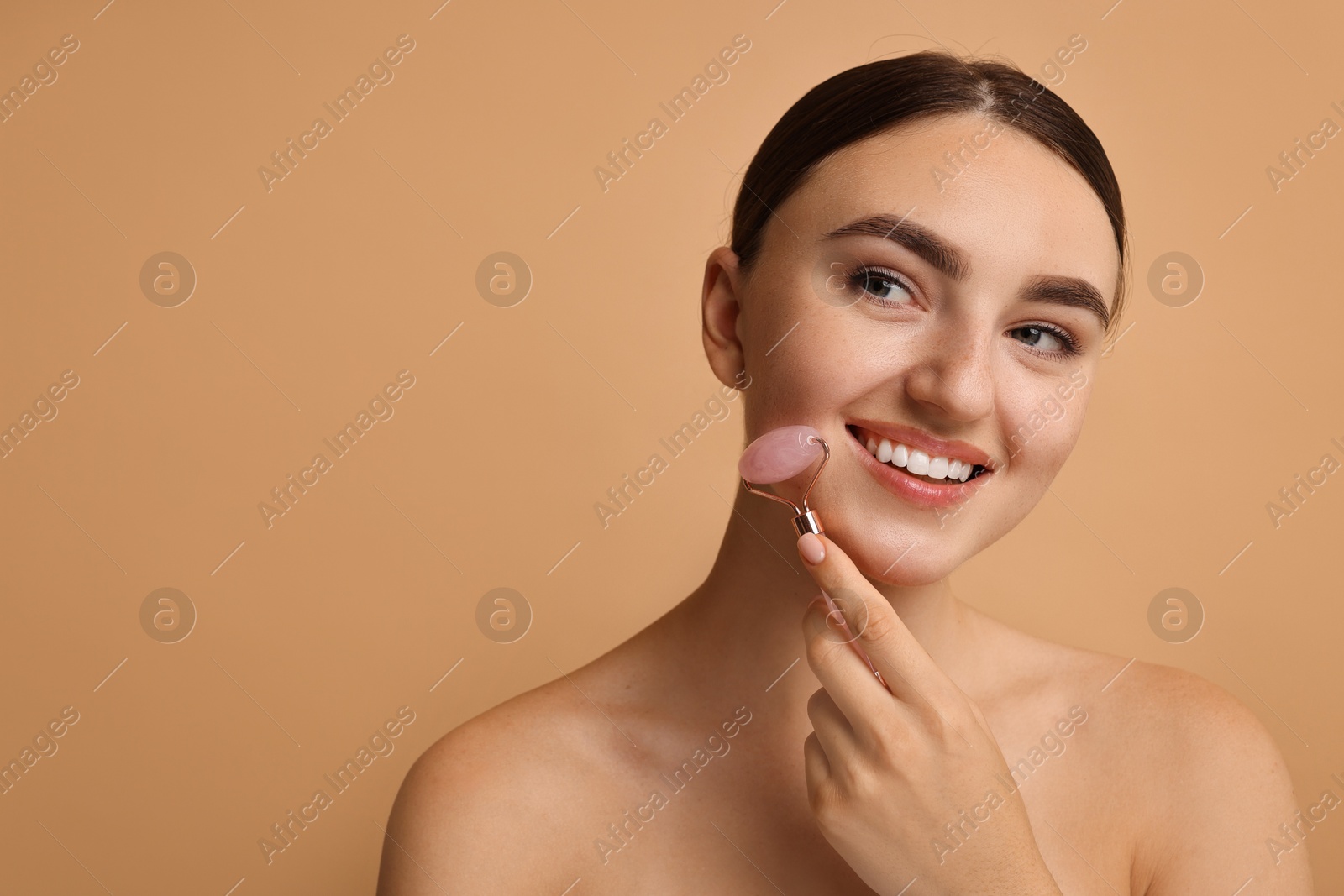 Photo of Beautiful young woman doing facial massage with roller on beige background, space for text