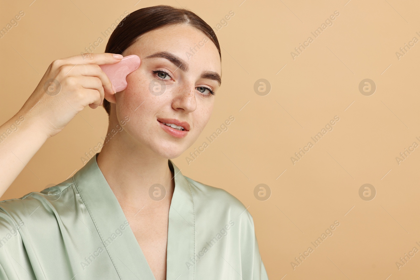 Photo of Beautiful young woman doing facial massage with gua sha tool on beige background, space for text