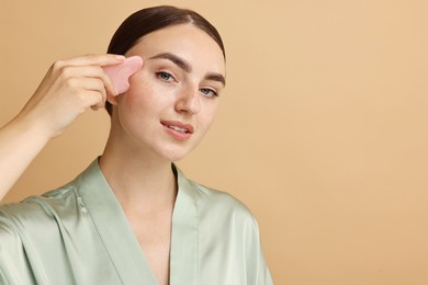 Photo of Beautiful young woman doing facial massage with gua sha tool on beige background, space for text