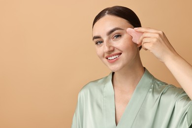 Beautiful young woman doing facial massage with gua sha tool on beige background, space for text