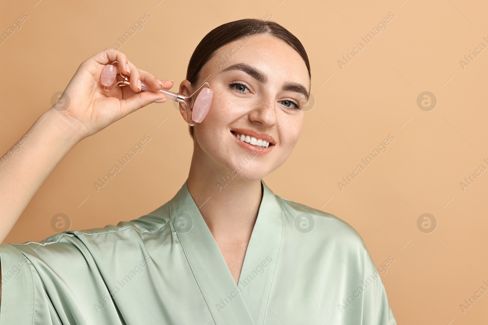 Photo of Beautiful young woman doing facial massage with roller on beige background