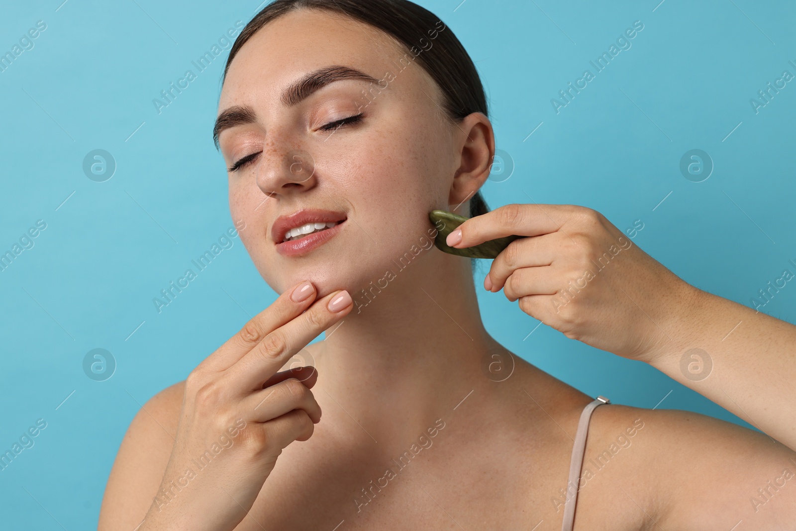 Photo of Beautiful young woman doing facial massage with gua sha tool on light blue background