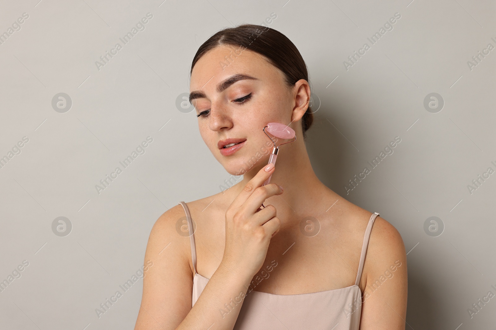 Photo of Beautiful young woman doing facial massage with roller on grey background