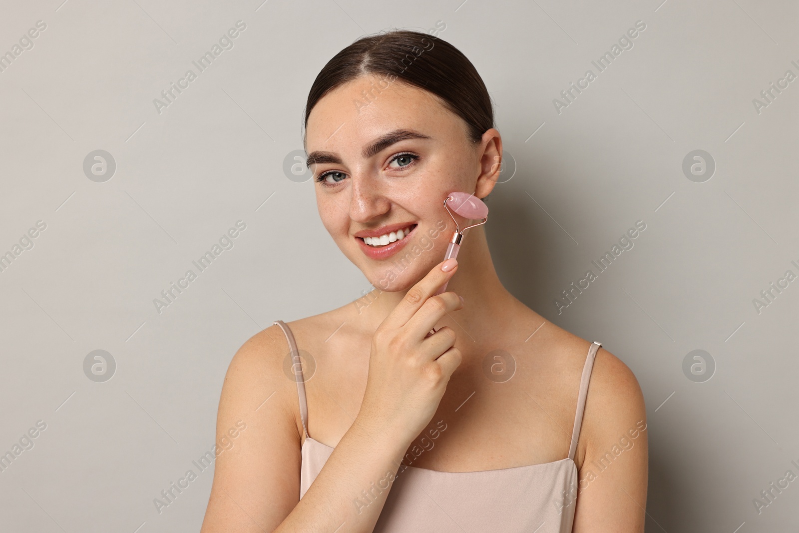 Photo of Beautiful young woman doing facial massage with roller on grey background