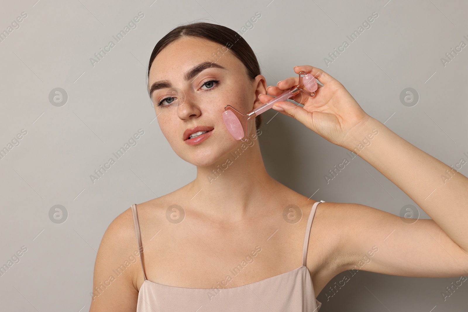 Photo of Beautiful young woman doing facial massage with roller on grey background