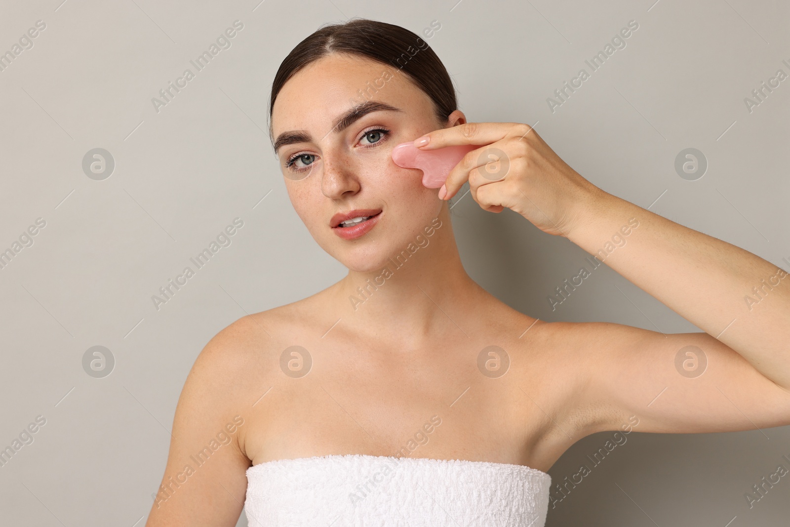 Photo of Beautiful young woman doing facial massage with gua sha tool on grey background