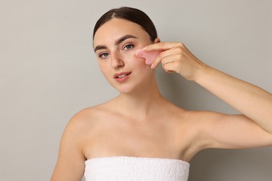 Beautiful young woman doing facial massage with gua sha tool on grey background