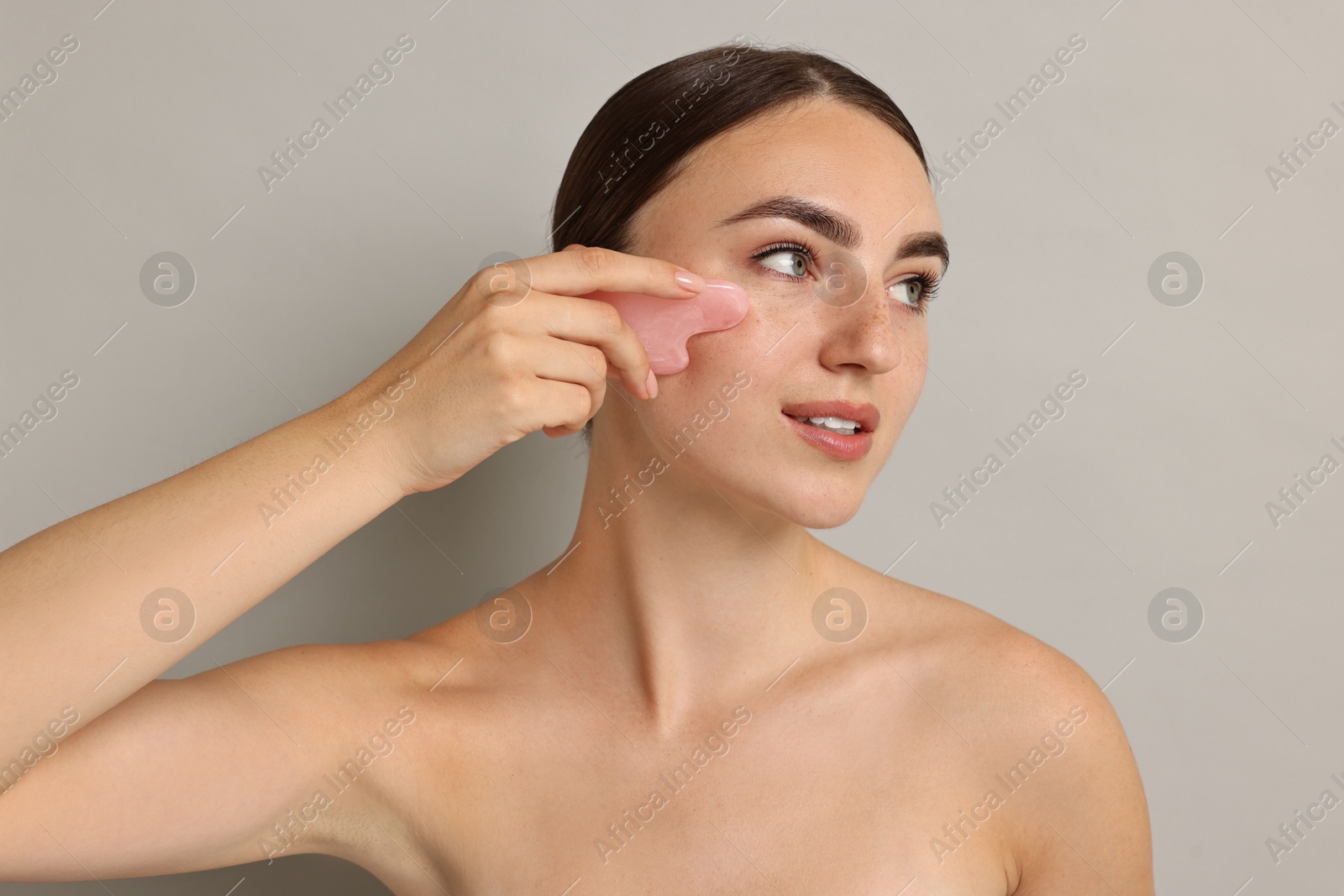 Photo of Beautiful young woman doing facial massage with gua sha tool on grey background