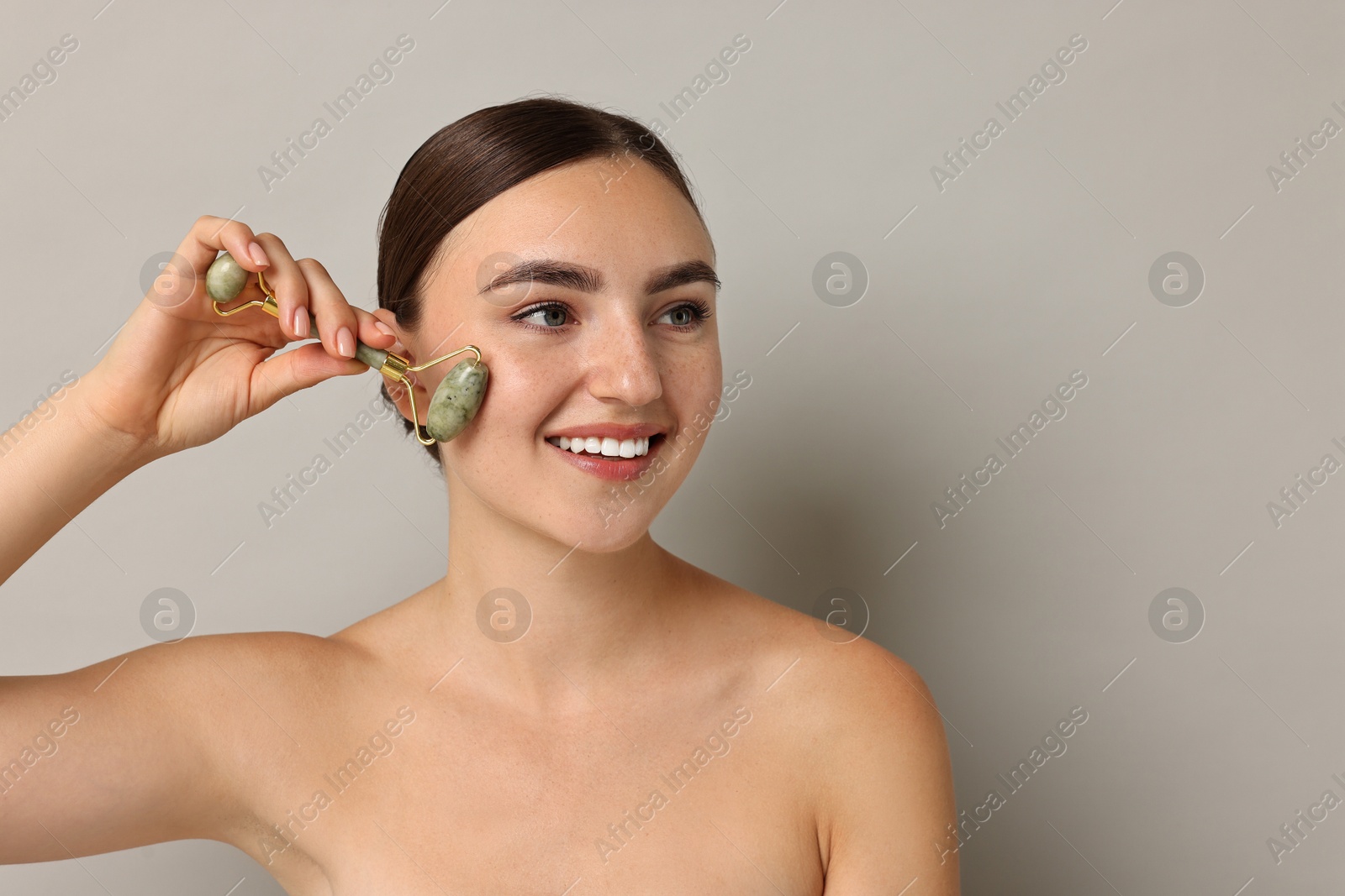 Photo of Beautiful young woman doing facial massage with roller on grey background, space for text