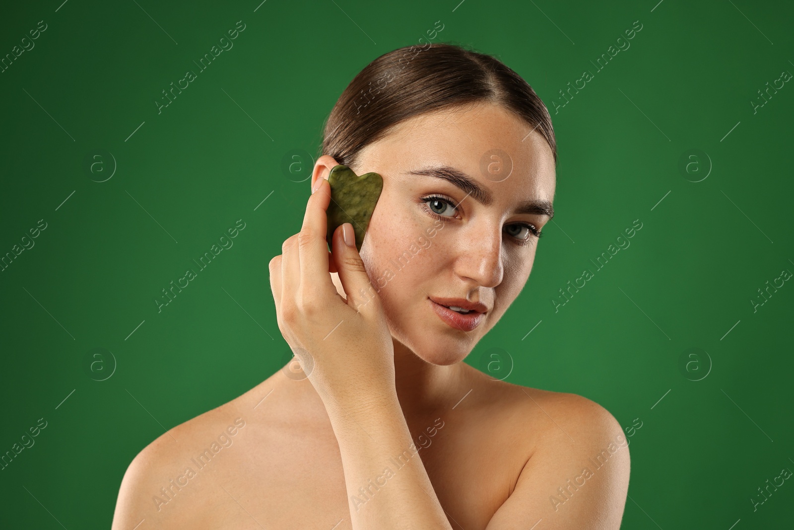 Photo of Beautiful young woman doing facial massage with gua sha tool on green background