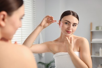 Photo of Beautiful young woman doing facial massage with roller near mirror at home