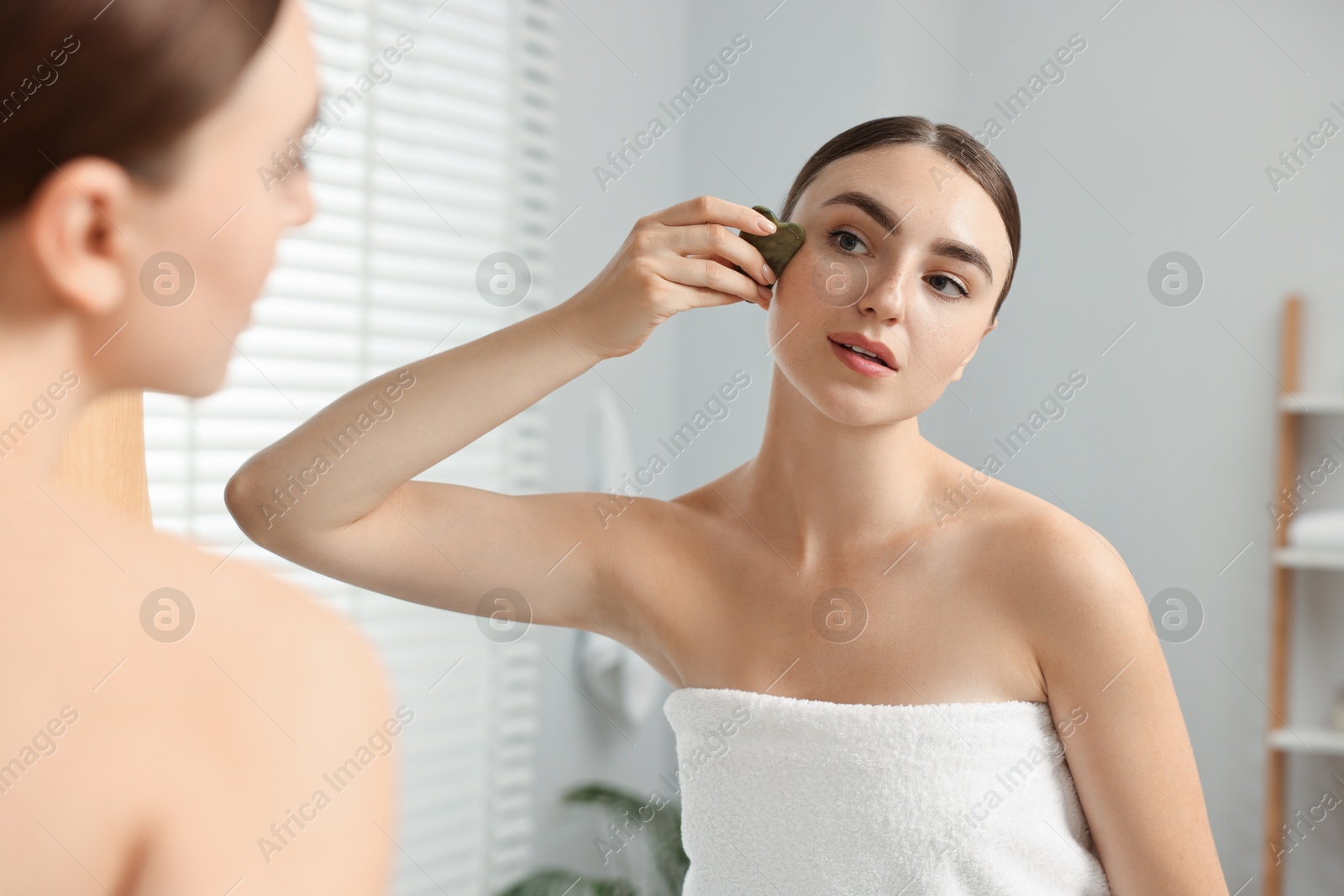 Photo of Beautiful young woman doing facial massage with gua sha tool near mirror at home