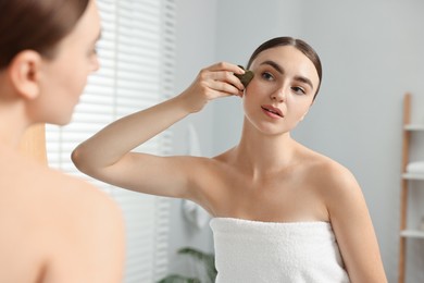 Beautiful young woman doing facial massage with gua sha tool near mirror at home