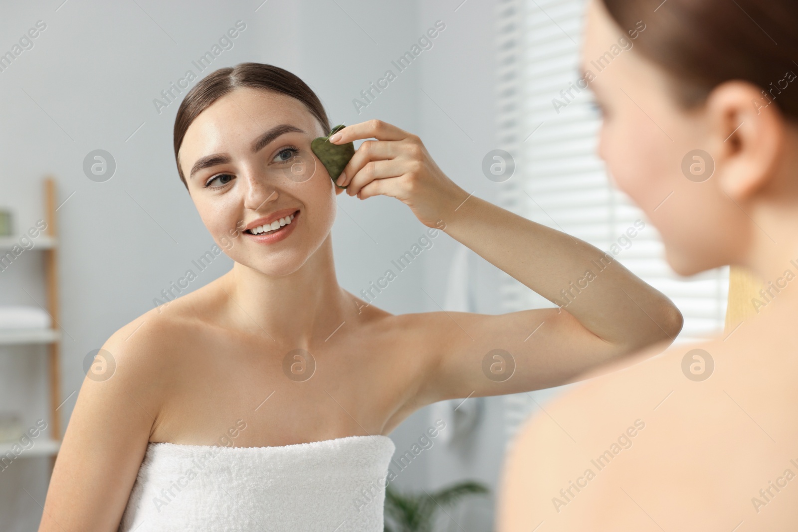 Photo of Beautiful young woman doing facial massage with gua sha tool near mirror at home