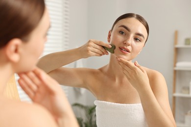 Photo of Beautiful young woman doing facial massage with gua sha tool near mirror at home