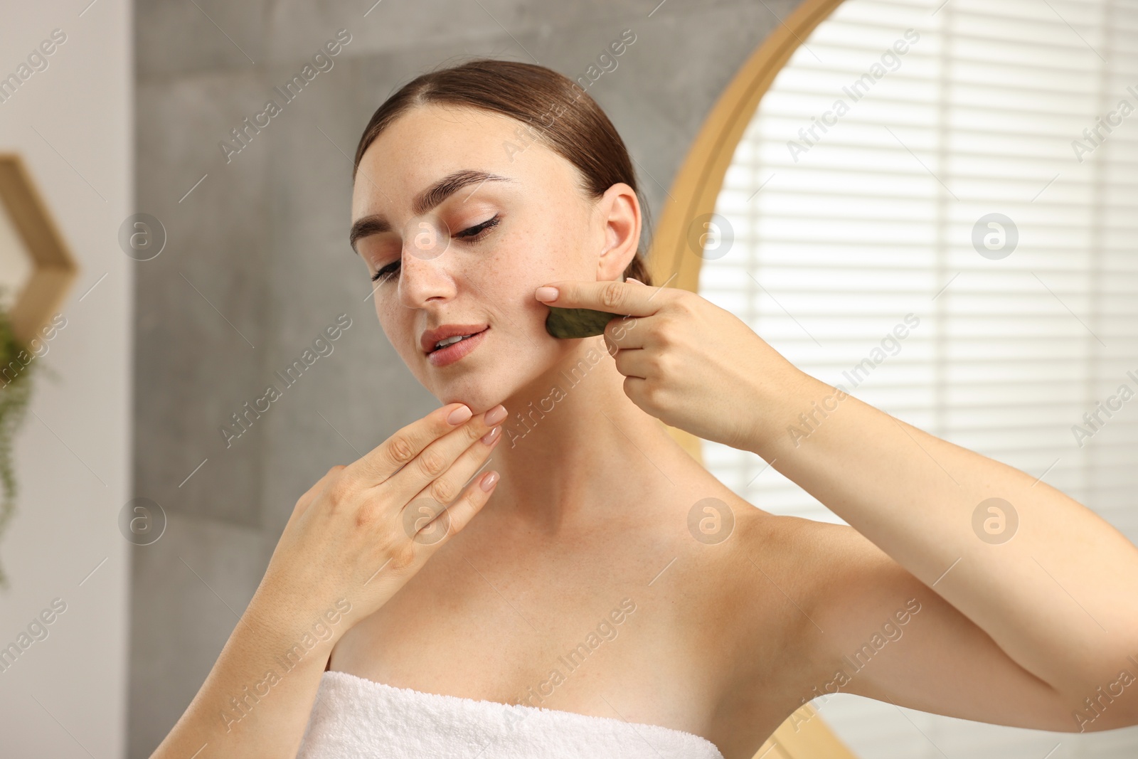 Photo of Beautiful young woman doing facial massage with gua sha tool indoors