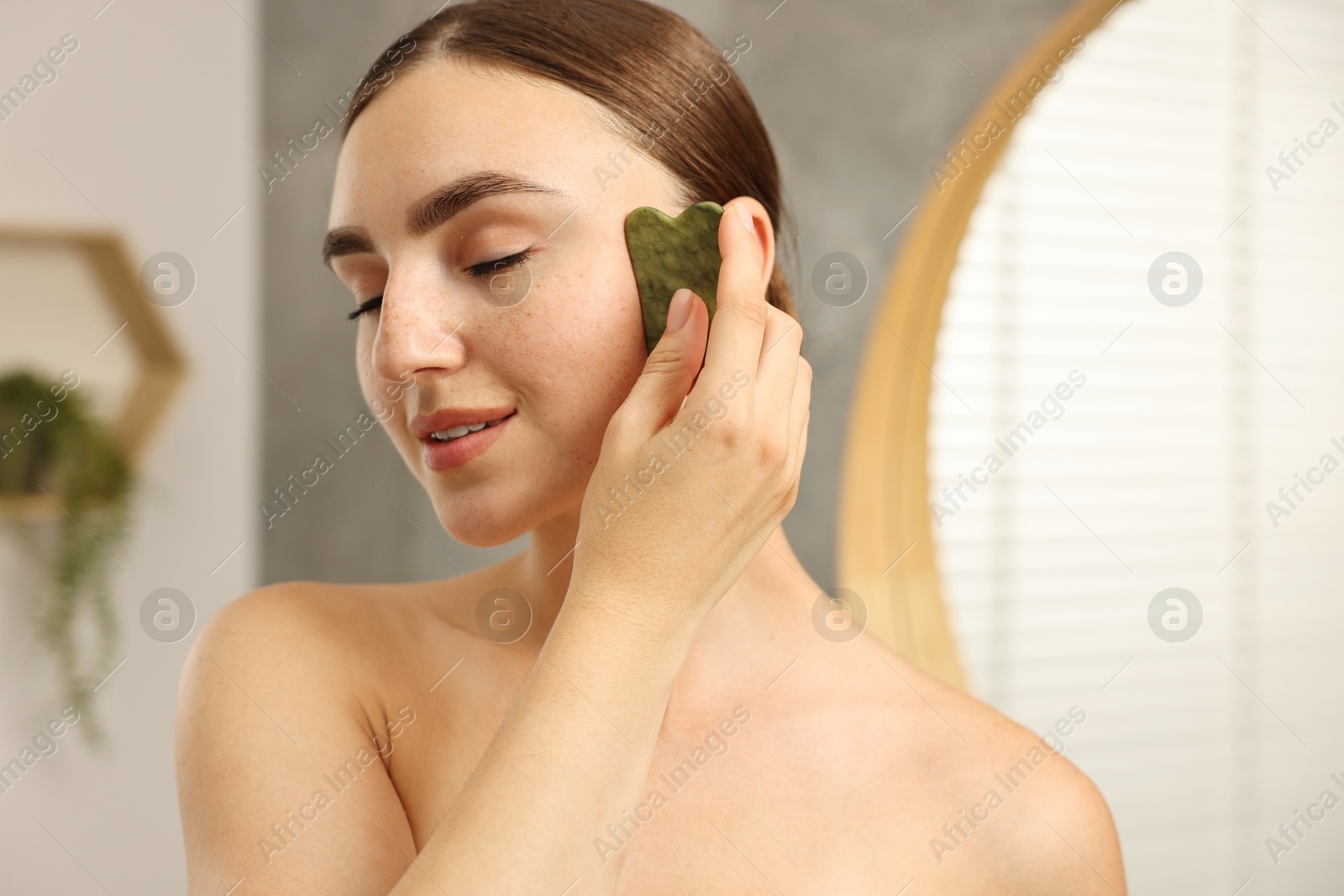 Photo of Beautiful young woman doing facial massage with gua sha tool indoors