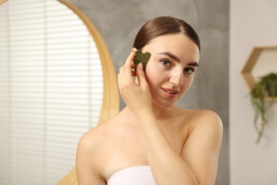 Photo of Beautiful young woman doing facial massage with gua sha tool indoors