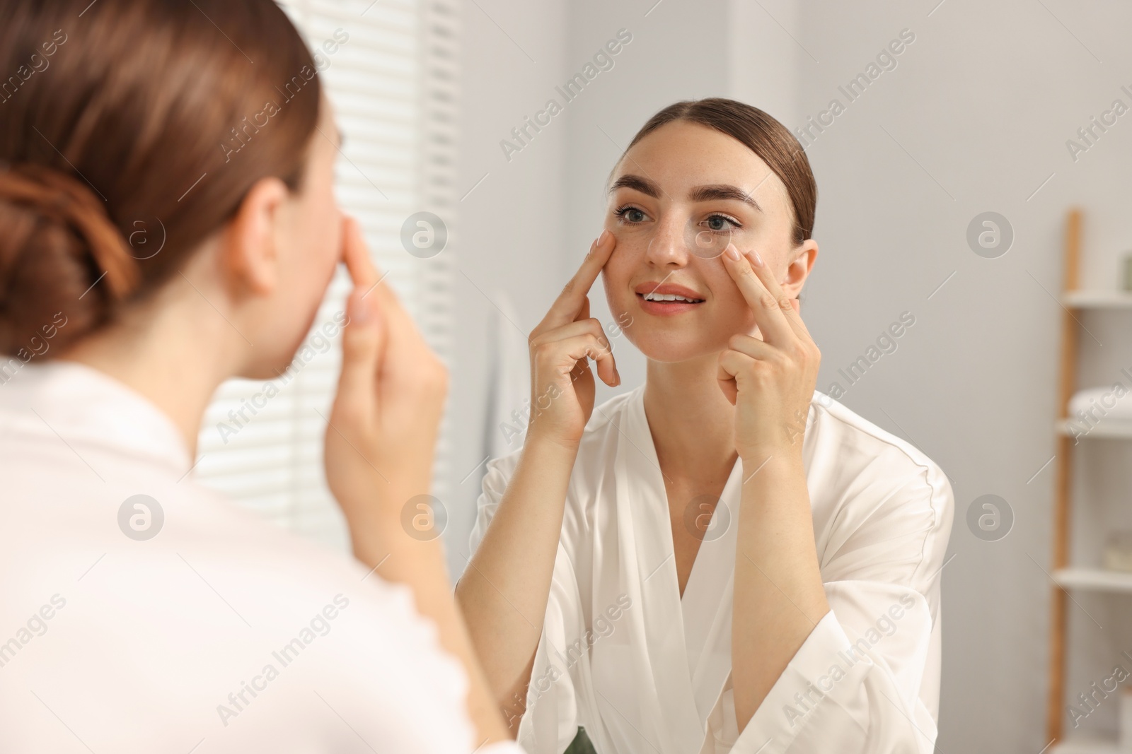 Photo of Beautiful young woman doing facial massage near mirror at home