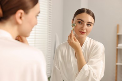 Beautiful young woman doing facial massage with roller near mirror at home