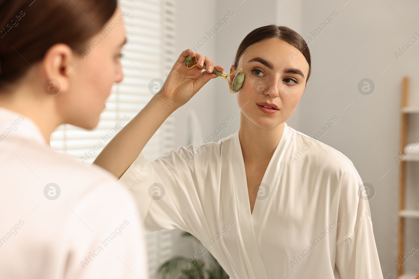 Photo of Beautiful young woman doing facial massage with roller near mirror at home