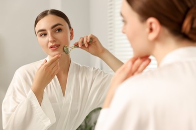 Photo of Beautiful young woman doing facial massage with roller near mirror at home