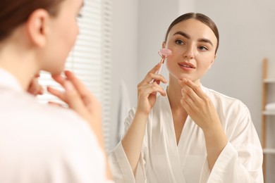 Photo of Beautiful young woman doing facial massage with roller near mirror at home