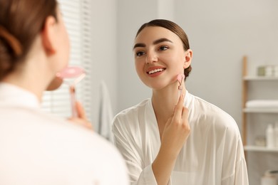 Photo of Beautiful young woman doing facial massage with roller near mirror at home