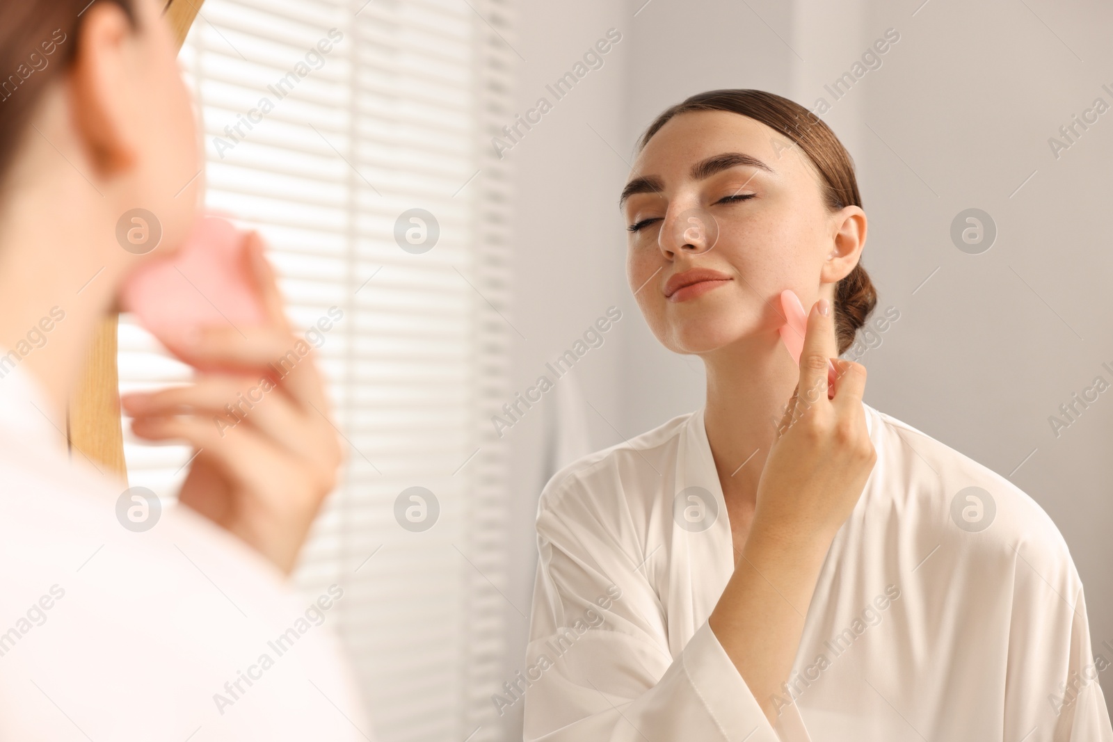 Photo of Beautiful young woman doing facial massage with gua sha tool near mirror at home