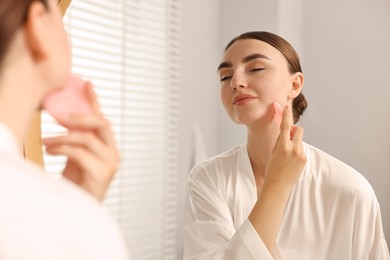 Photo of Beautiful young woman doing facial massage with gua sha tool near mirror at home