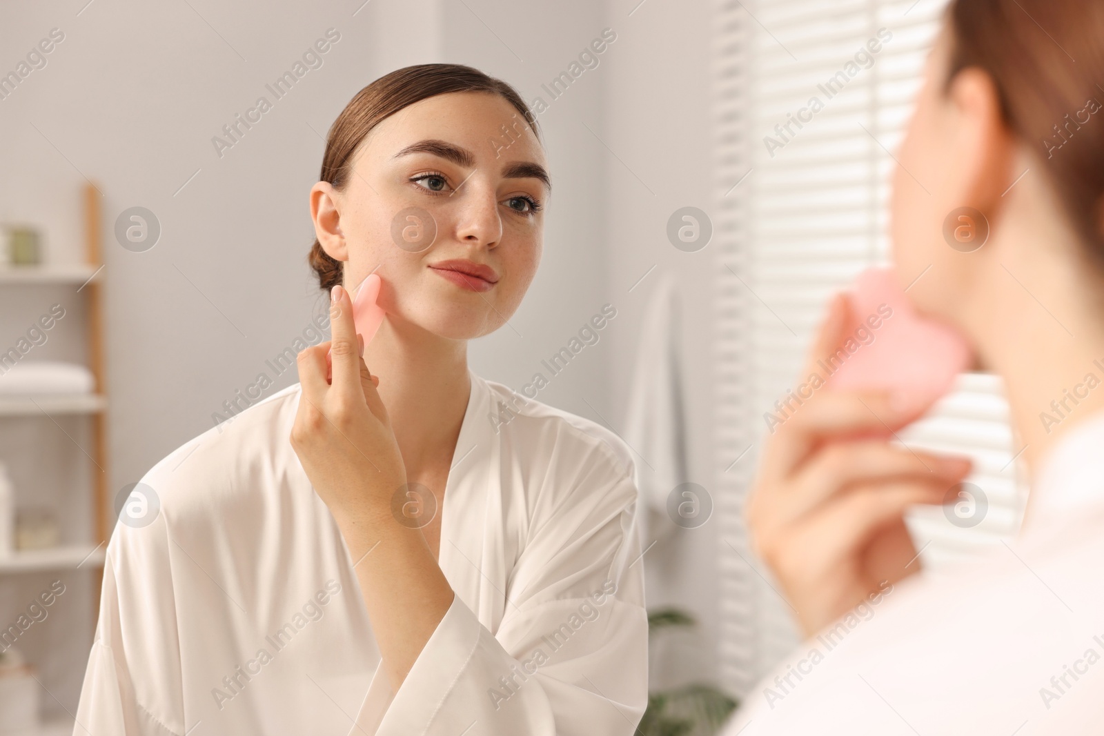 Photo of Beautiful young woman doing facial massage with gua sha tool near mirror at home