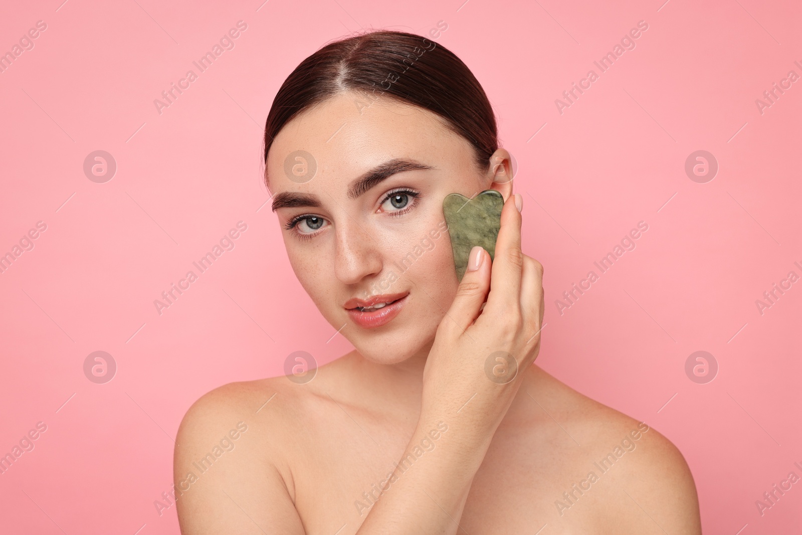 Photo of Beautiful young woman doing facial massage with gua sha tool on pink background