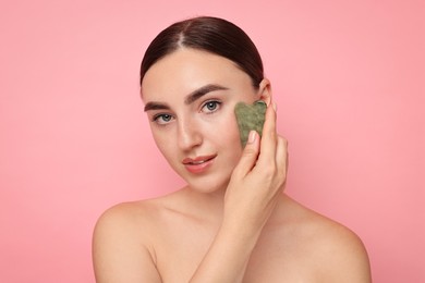 Beautiful young woman doing facial massage with gua sha tool on pink background