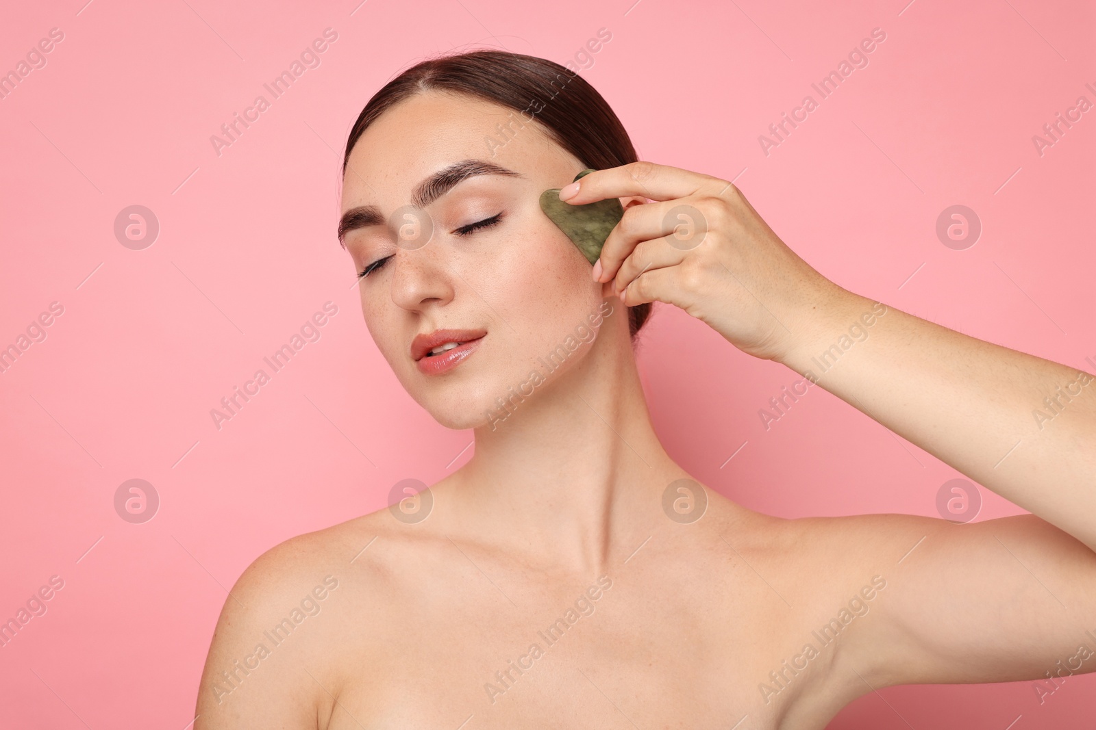 Photo of Beautiful young woman doing facial massage with gua sha tool on pink background