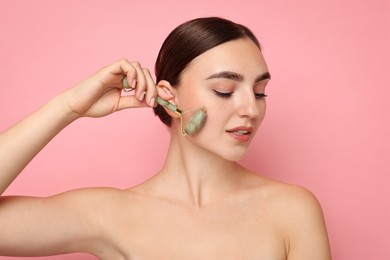 Beautiful young woman doing facial massage with roller on pink background