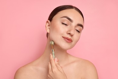 Beautiful young woman doing facial massage with roller on pink background