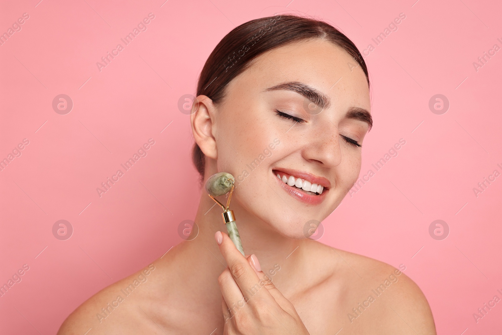 Photo of Beautiful young woman doing facial massage with roller on pink background