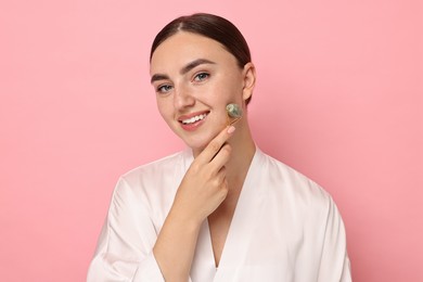 Beautiful young woman doing facial massage with roller on pink background