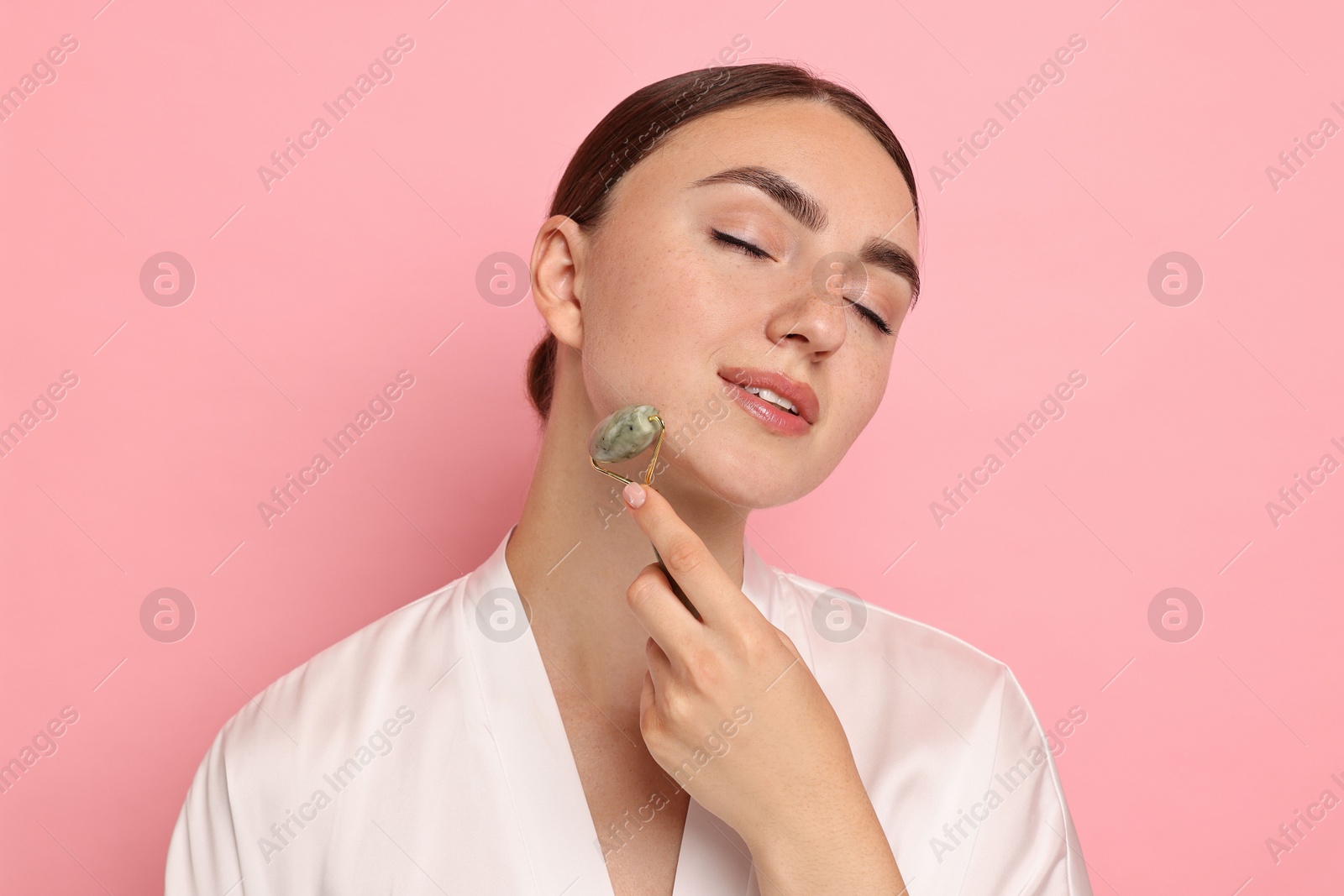 Photo of Beautiful young woman doing facial massage with roller on pink background