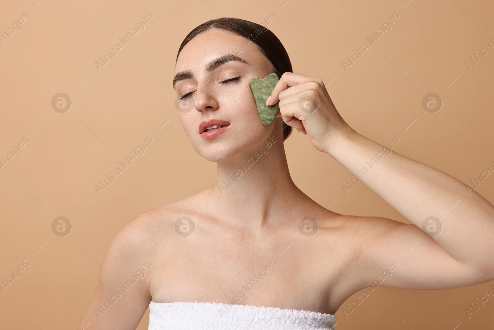 Photo of Beautiful young woman doing facial massage with gua sha tool on beige background