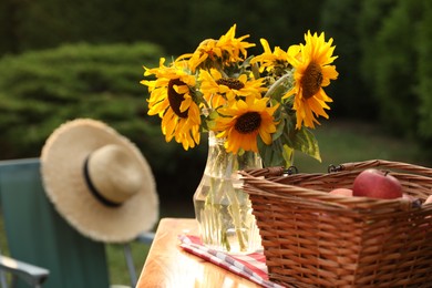 Vase of sunflowers and wicker basket with apples on wooden table in garden