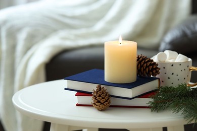 Photo of Burning candle, books, fir branches and cones on coffee table indoors. Christmas atmosphere