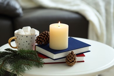 Photo of Burning candle, books, fir branches and cones on coffee table indoors. Christmas atmosphere