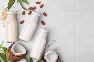 Photo of Different types of vegan milk in bottles with ingredients and green leaves on white tiled table, flat lay. Space for text