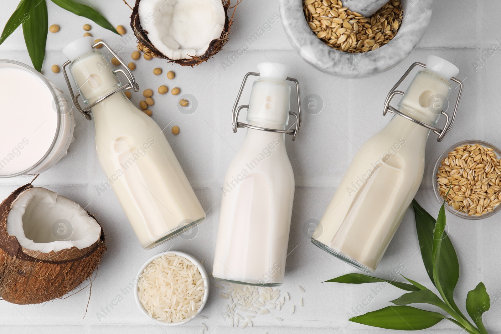 Photo of Different types of vegan milk in bottles with ingredients and green leaves on white tiled table, flat lay