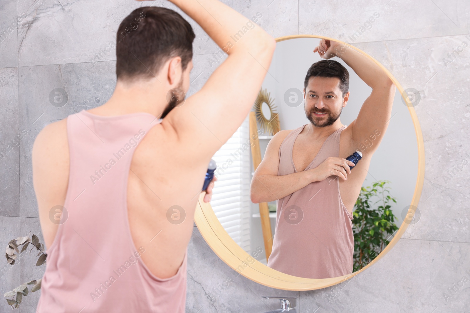 Photo of Smiling man applying roll-on deodorant near mirror at home, back view