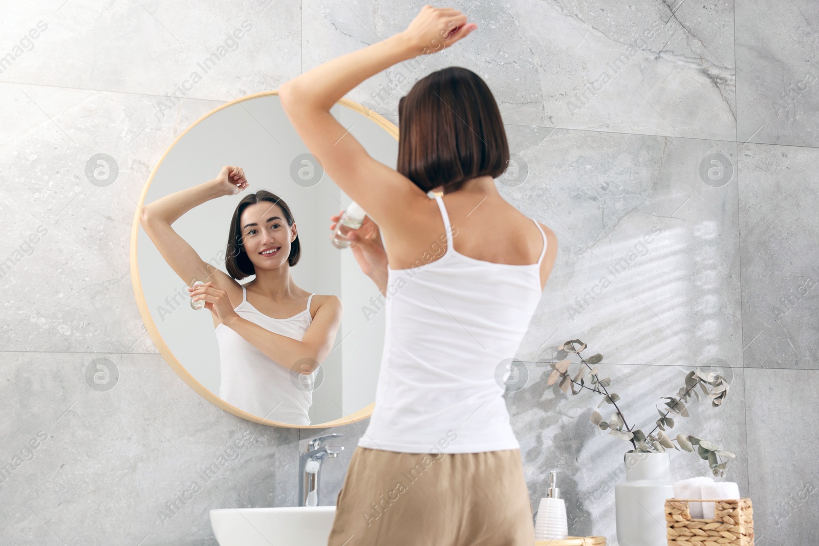 Photo of Smiling woman applying roll-on deodorant near mirror at home, back view