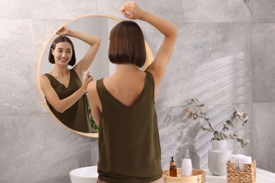 Smiling woman applying spray deodorant near mirror at home, back view