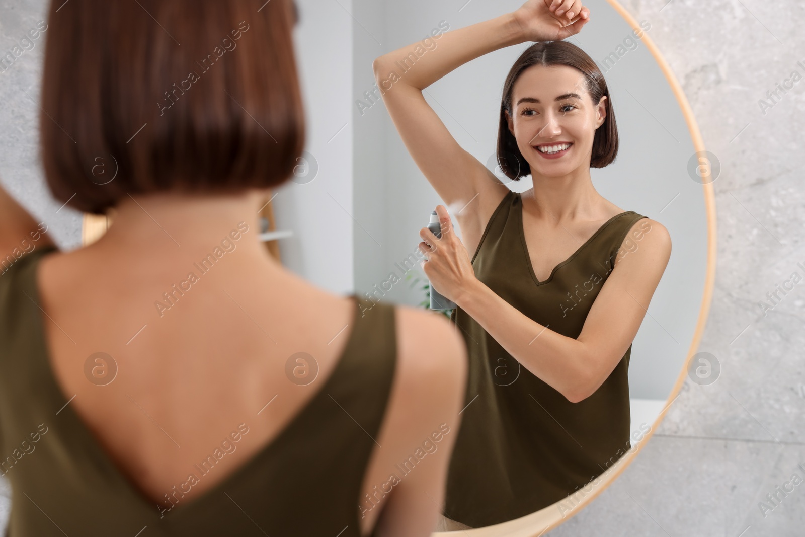 Photo of Smiling woman applying spray deodorant near mirror at home, back view