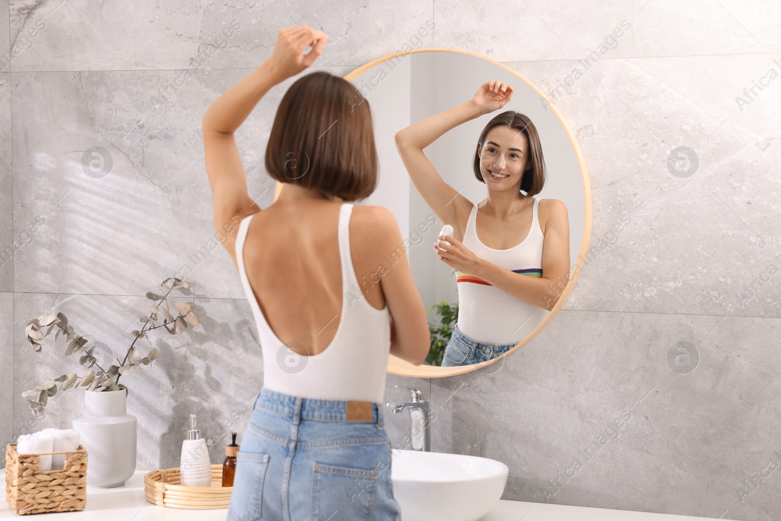 Photo of Smiling woman applying roll-on deodorant near mirror at home, back view